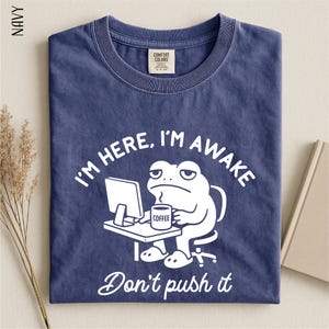 May include: Navy blue t-shirt with a white graphic of a frog at a desk, holding a coffee mug. The text reads "I'M HERE, I'M AWAKE" and "Don't push it". The shirt has a crew neck and a Comfort Colors tag.