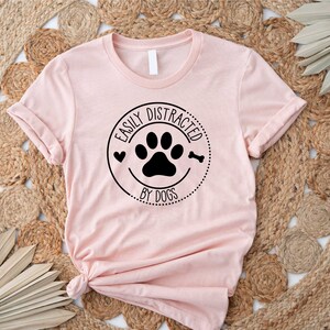 May include: A pink t-shirt with a black circle graphic that says "Easily Distracted by Dogs" with a paw print and a bone inside the circle.