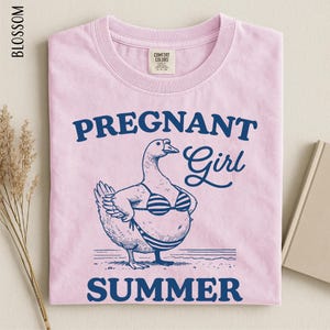 A pink t-shirt with a graphic design depicting a cartoon duck wearing a bikini top and the text "Pregnant Girl Summer" printed on it.