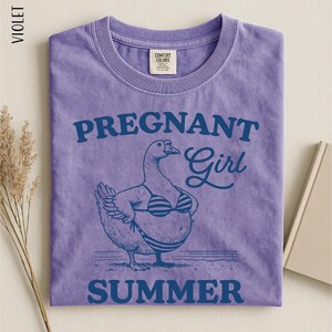A purple t-shirt with a graphic design depicting a cartoon duck wearing a bikini top and the text "Pregnant Girl Summer" printed on it.