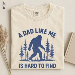 A t-shirt with a graphic design depicting a blue Sasquatch silhouette in a forest setting, accompanied by the text "A DAD LIKE ME IS HARD TO FIND".
