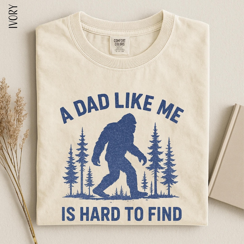 A t-shirt with a graphic design depicting a blue Sasquatch silhouette in a forest setting, accompanied by the text "A DAD LIKE ME IS HARD TO FIND".