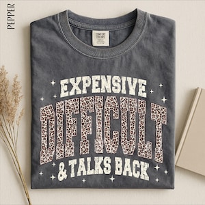 May include: A charcoal gray crew neck t-shirt featuring the text "EXPENSIVE DIFFICULT & TALKS BACK" in a distressed white font. The word "DIFFICULT" is filled with a brown and black leopard print pattern, surrounded by small white star icons.