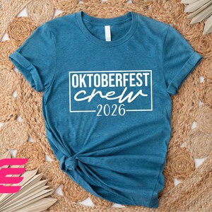 A blue t-shirt with the text "OKTOBERFEST CREW 2026" printed on it, along with a graphic of a person wearing a t-shirt. The t-shirt is placed on a woven mat, and there are some pink and brown circular shapes in the background.