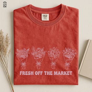 a red t-shirt with a graphic design of four bouquets of flowers, accompanied by the text "FRESH OFF THE MARKET" printed on it.