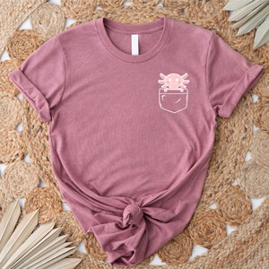 Pu&ograve; includere: Una maglietta color malva con una tasca bianca che presenta un axolotl dei cartoni animati. L'axolotl &egrave; rosa con un contorno bianco e ha un viso sorridente.