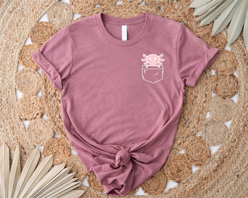 Pu&ograve; includere: Una maglietta color malva con una tasca bianca che presenta un axolotl dei cartoni animati. L'axolotl &egrave; rosa con un contorno bianco e ha un viso sorridente.