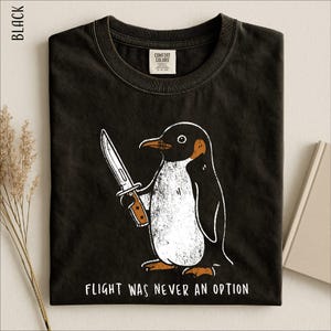 May include: A black crew neck t-shirt featuring a graphic of a penguin holding a knife. The text below the penguin reads, "FLIGHT WAS NEVER AN OPTION." The shirt has a Comfort Colors brand label on the inside of the collar and a black color label.