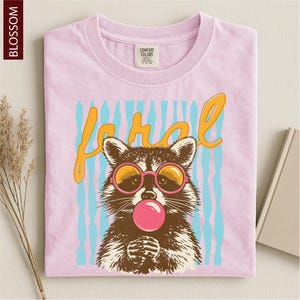 A pink t-shirt with a graphic design of a raccoon wearing sunglasses and blowing a bubble gum bubble.