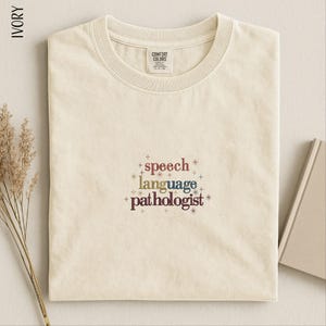 May include: An ivory-colored crew neck t-shirt featuring the embroidered text "speech language pathologist" in a multi-colored font, surrounded by small starburst icons. The shirt has a Comfort Colors brand label on the inside of the collar.