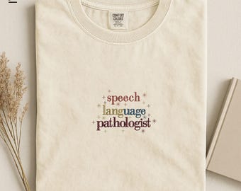 May include: An ivory-colored crew neck t-shirt featuring the embroidered text "speech language pathologist" in a multi-colored font, surrounded by small starburst icons. The shirt has a Comfort Colors brand label on the inside of the collar.