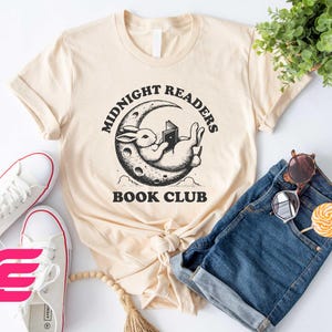 May include: A beige t-shirt with a black and white graphic of a rabbit reading a book on a crescent moon. The text "Midnight Readers Book Club" is printed on the shirt.