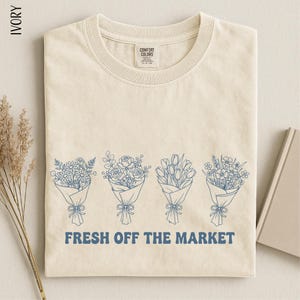 A beige t-shirt with a graphic design of four flower bouquets and the text "FRESH OFF THE MARKET" printed on it.