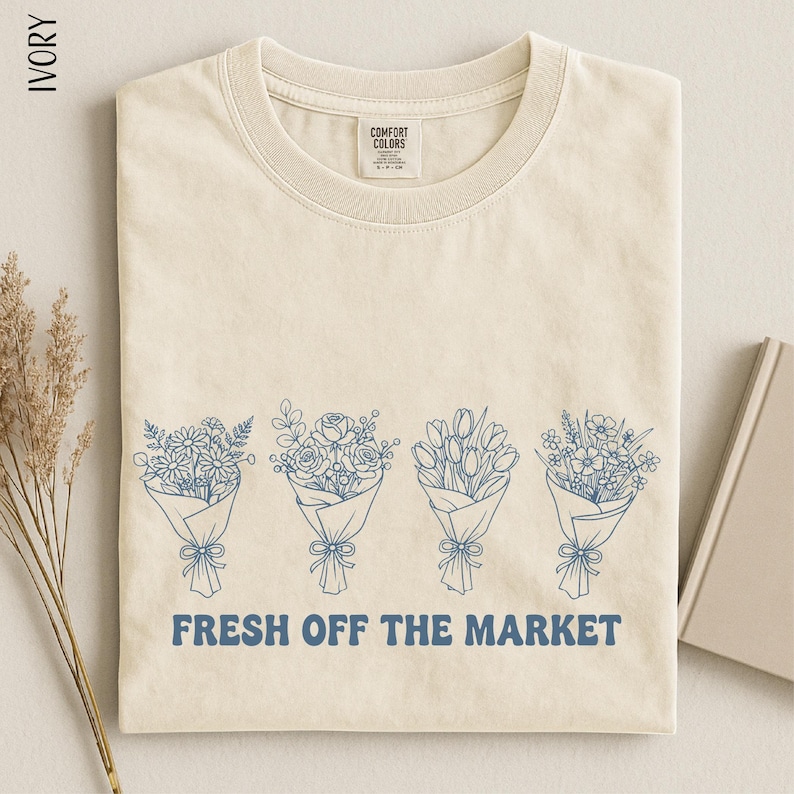 A beige t-shirt with a graphic design of four flower bouquets and the text "FRESH OFF THE MARKET" printed on it.