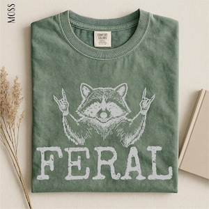 a green t-shirt with a graphic design of a raccoon making a rock and roll sign, accompanied by the word "FERAL" printed below.