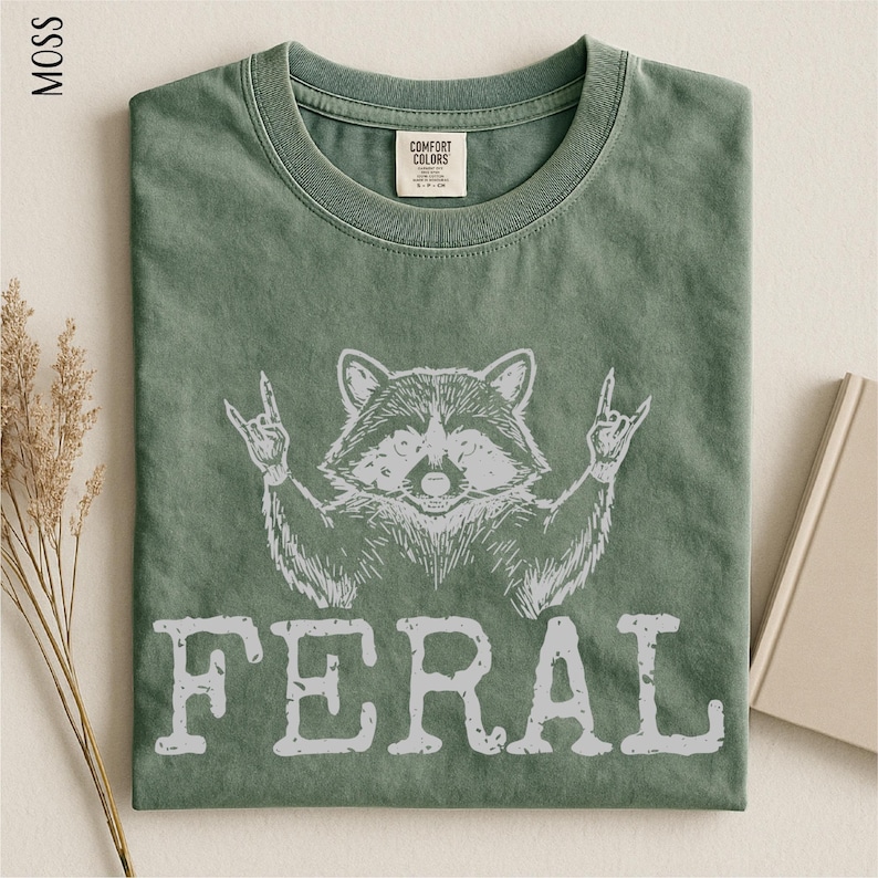 a green t-shirt with a graphic design of a raccoon making a rock and roll sign, accompanied by the word "FERAL" printed below.