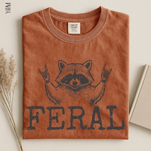 a brown t-shirt with a graphic design of a raccoon making a hand gesture, along with the word "FERAL" printed below it.