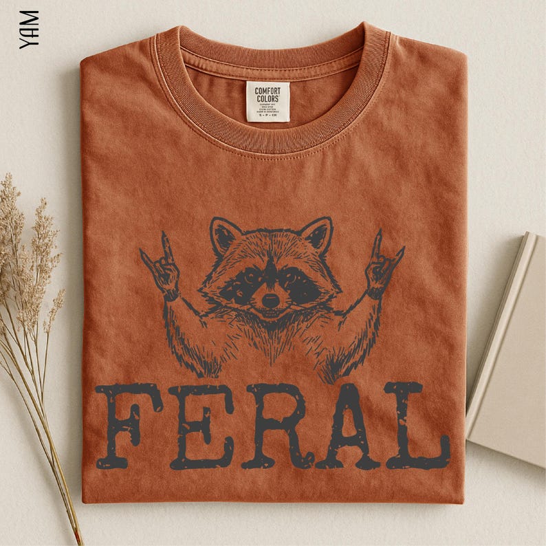 a brown t-shirt with a graphic design of a raccoon making a hand gesture, along with the word "FERAL" printed below it.