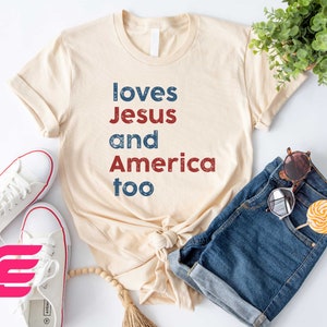 May include: A beige t-shirt with the text "loves Jesus and America too" in red, blue, and white lettering. The shirt is laid out on a white surface with a pair of white sneakers, a pair of blue denim shorts, and a pair of sunglasses.