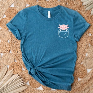 Pu&ograve; includere: Una t-shirt blu turchese con una tasca bianca che presenta un'illustrazione di axolotl rosa. La t-shirt &egrave; realizzata in un materiale morbido e confortevole.
