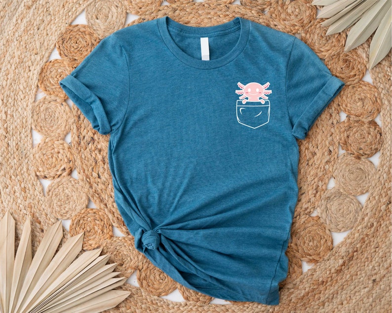 Pu&ograve; includere: Una t-shirt blu turchese con una tasca bianca che presenta un'illustrazione di axolotl rosa. La t-shirt &egrave; realizzata in un materiale morbido e confortevole.