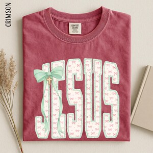 A red t-shirt with the word "JESUS" printed on it, along with a green ribbon bow. The t-shirt is folded and placed on a surface, with a book and some dried flowers nearby.