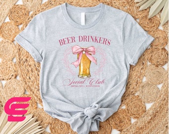 Puede incluir: Camiseta gris con un lazo rosa y un vaso de cerveza. El texto de la camiseta dice "Beer Drinkers Social Club Drinks Well With Others".