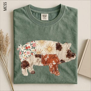 May include: A moss green t-shirt featuring a pig-shaped graphic composed of various fabric patches. The patches include floral patterns in red and white, sunburst designs, and brown cow-print textures. The shirt has a crew neckline and a label.