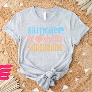 May include: Gray t-shirt with the text "Saltwater Seashells Sunshine" in blue, pink, and yellow. The text is stylized with a wave, seashell, and sun graphic.