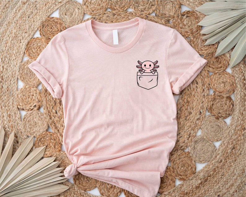 Pu&ograve; includere: Una maglietta rosa con una tasca che presenta un axolotl dei cartoni animati. L'axolotl &egrave; rosa con occhi neri e pinne.