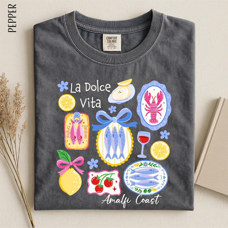 a gray t-shirt with a vibrant graphic design depicting various food items and icons, including fish, lemons, and a lobster, along with the text "La Dolce Vita" and "Amalfi Coast".