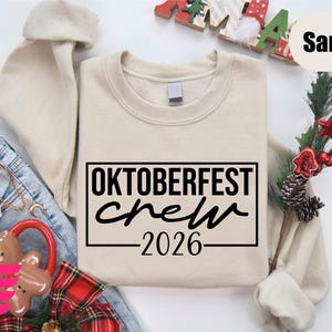 a beige sweatshirt with the text "OKTOBERFEST CREW 2026" printed on it, along with some other clothing items and holiday decorations.