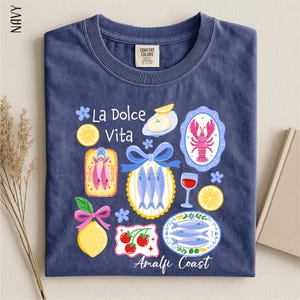A navy blue t-shirt with a colorful graphic design depicting various food items and icons, including fish, lemons, and a lobster, along with the text "La Dolce Vita" and "Amalfi Coast".