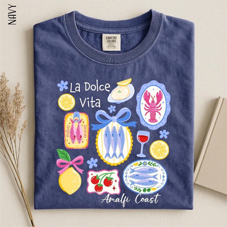 A navy blue t-shirt with a colorful graphic design depicting various food items and icons, including fish, lemons, and a lobster, along with the text "La Dolce Vita" and "Amalfi Coast".