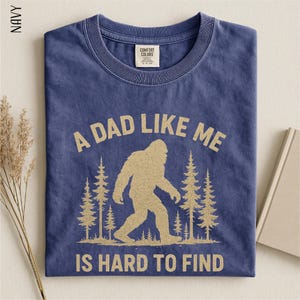 A navy blue t-shirt with a graphic design of a Sasquatch silhouette and the text "A DAD LIKE ME IS HARD TO FIND" printed on it.