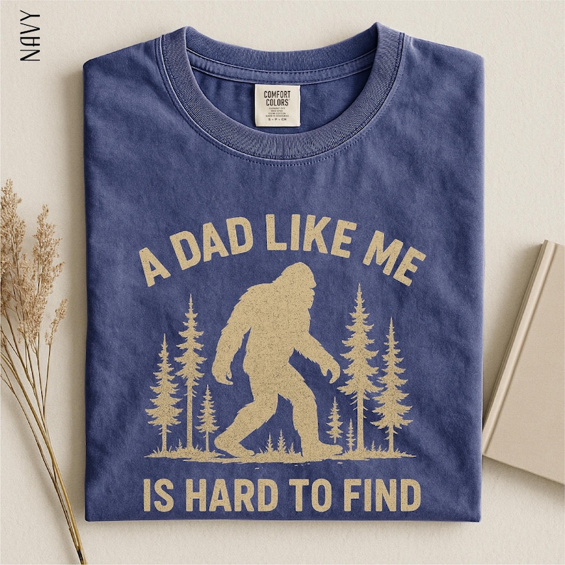 A navy blue t-shirt with a graphic design of a Sasquatch silhouette and the text "A DAD LIKE ME IS HARD TO FIND" printed on it.