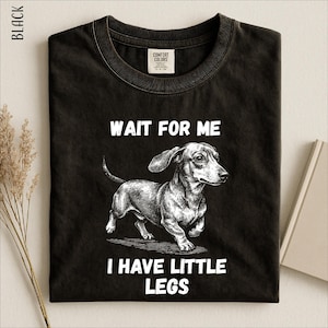 A black t-shirt with a graphic design of a dachshund dog and the text "WAIT FOR ME I HAVE LITTLE LEGS" printed on it.