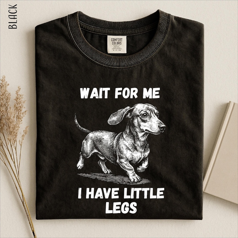 A black t-shirt with a graphic design of a dachshund dog and the text "WAIT FOR ME I HAVE LITTLE LEGS" printed on it.
