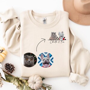 K&ouml;nnte beinhalten: Ein beiger Pullover mit einem Design aus zwei Haustieren, einer Katze und einem Hund, in einem Kreis auf der Vorderseite. Der Text "Chico & Fox" ist unter dem Design gedruckt.