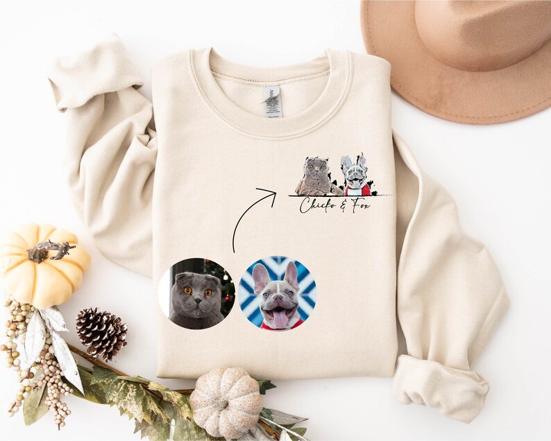K&ouml;nnte beinhalten: Ein beiger Pullover mit einem Design aus zwei Haustieren, einer Katze und einem Hund, in einem Kreis auf der Vorderseite. Der Text "Chico & Fox" ist unter dem Design gedruckt.