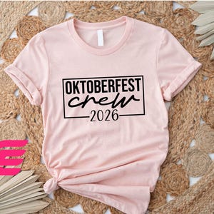 A pink t-shirt with the text "OKTOBERfest crew 2026" printed on it, along with a graphic of a pink flower.