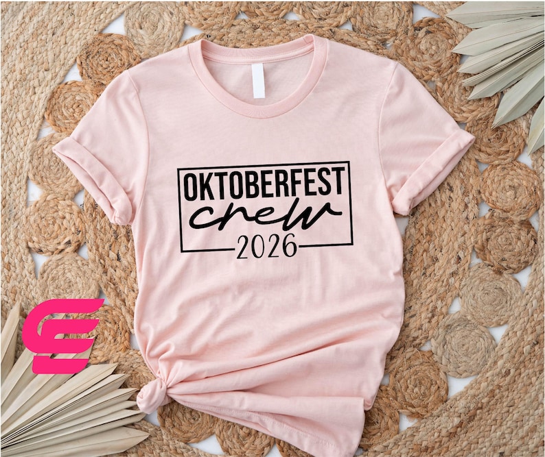 A pink t-shirt with the text "OKTOBERfest crew 2026" printed on it, along with a graphic of a pink flower.