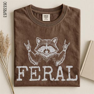 a brown t-shirt with a graphic design of a raccoon making a rock and roll sign, accompanied by the word "FERAL" printed below.