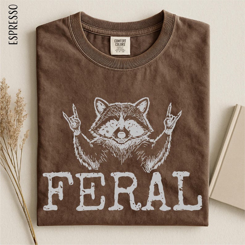 a brown t-shirt with a graphic design of a raccoon making a rock and roll sign, accompanied by the word "FERAL" printed below.