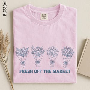 A pink t-shirt with a graphic design of four bouquets of flowers and the text "FRESH OFF THE MARKET" printed on it.