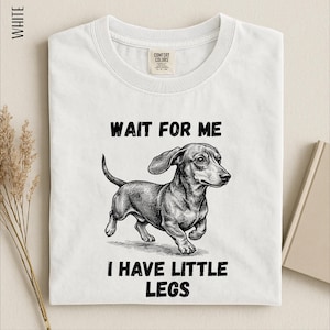 A white t-shirt with a black and white illustration of a dachshund dog and text that reads "WAIT FOR ME I HAVE LITTLE LEGS".