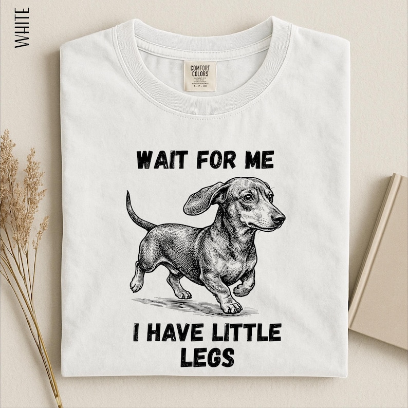 A white t-shirt with a black and white illustration of a dachshund dog and text that reads "WAIT FOR ME I HAVE LITTLE LEGS".
