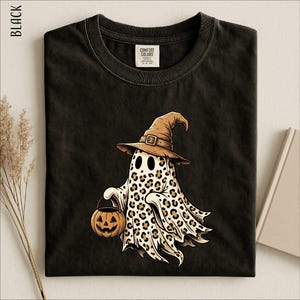 May include: A black t-shirt featuring a graphic of a ghost wearing a brown witch hat and holding a small orange jack-o'-lantern. The ghost has a tan and brown leopard print pattern across its body. The shirt has a Comfort Colors brand label.