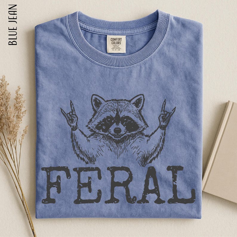 A blue t-shirt with a graphic design of a raccoon making a peace sign and the word "FERAL" below it.