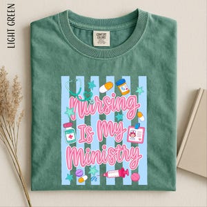 May include: A light green t-shirt featuring a graphic with light blue vertical stripes and the text "Nursing Is My Ministry" in pink. The design includes illustrations of a stethoscope, pills, a medicine bottle, and a nurse identification badge.